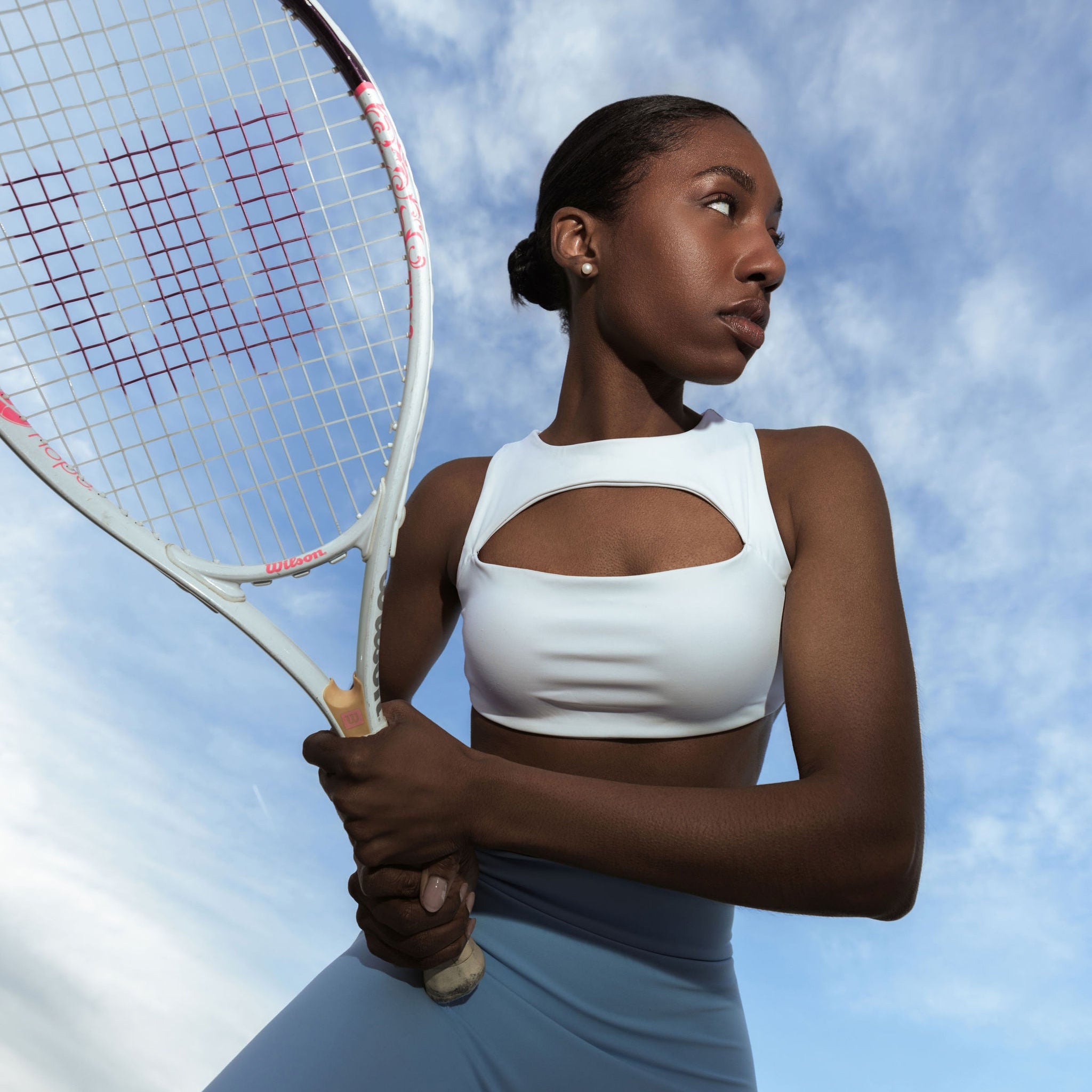 Model in The Palm activewear set with tennis racket and sky background. Clean aesthetic.