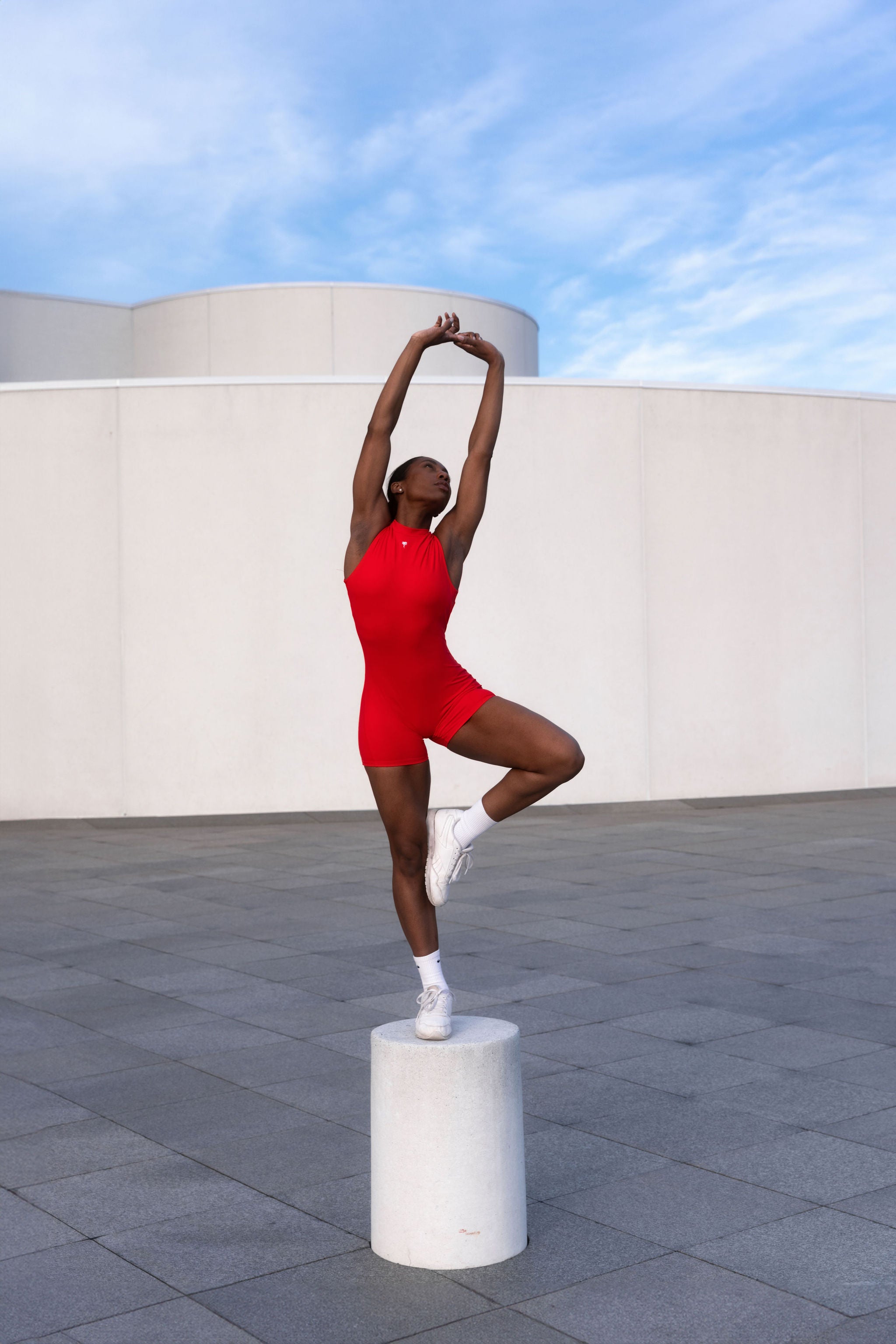 Model in sculpting red romper standing in ballet pose under blue sky — The Palm activewear