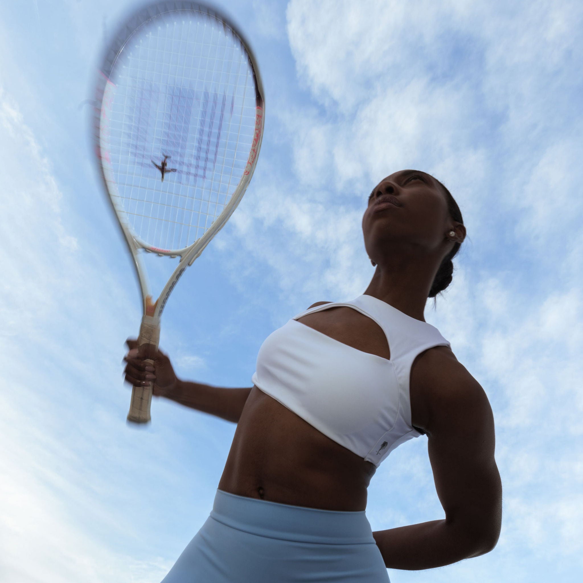 White cut-out sports bra and blue leggings by The Palm, worn by athletic woman with tennis racket under the sky.