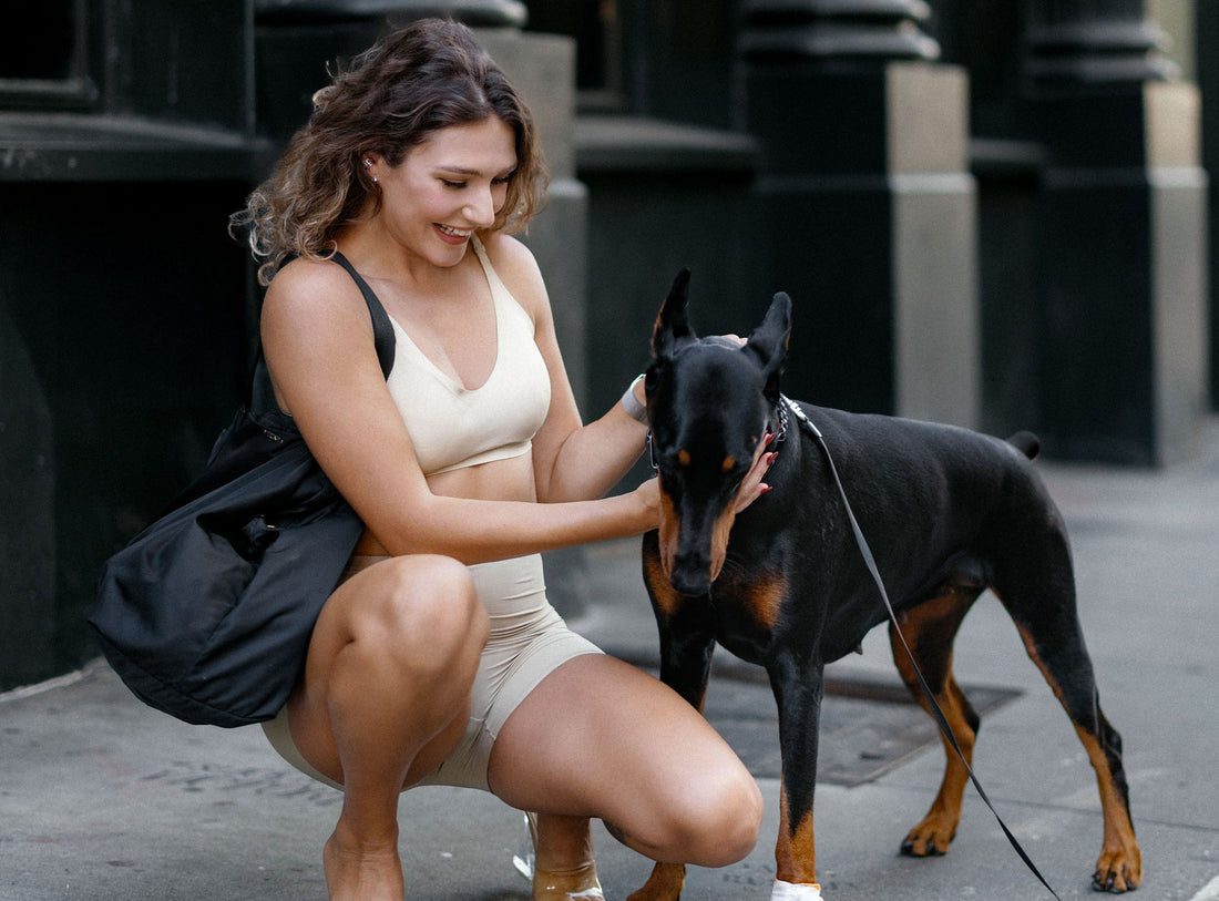A woman in cream-colored athletic wear crouching on a city sidewalk and smiling while petting her black Doberman dog.