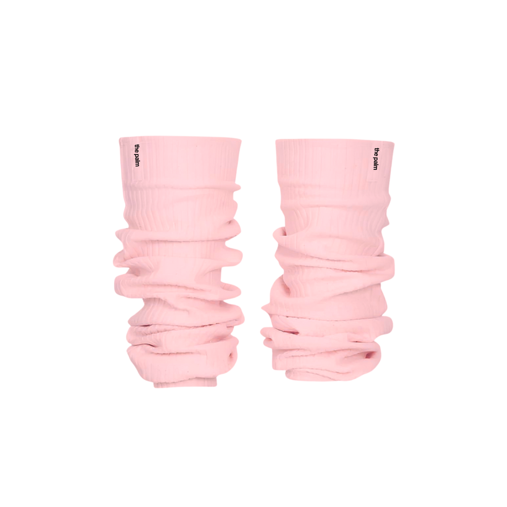 Ivory ribbed knit leg warmers by The Palm, featuring a minimalist logo tag and soft slouchy texture on a powder pink background.1
