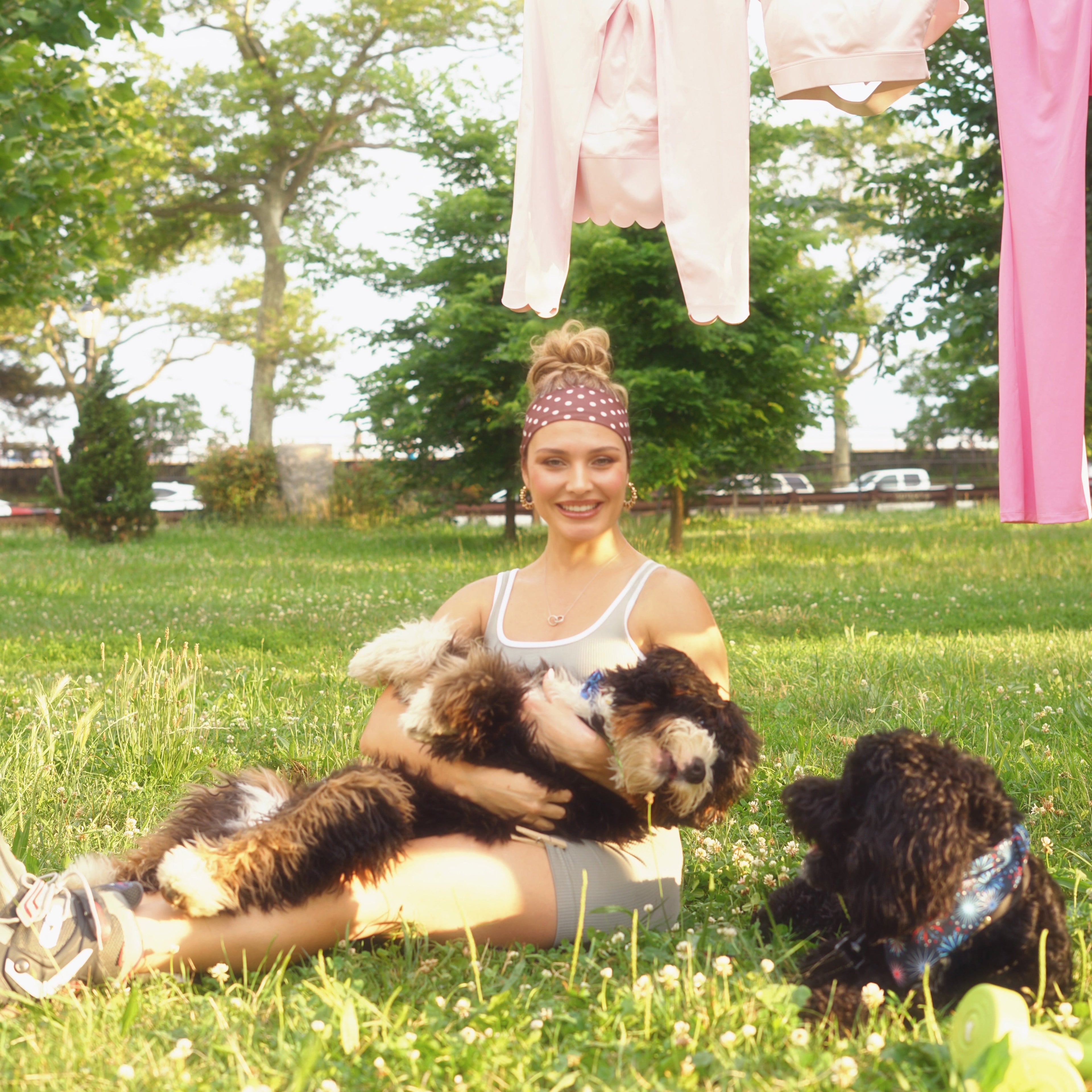 A woman sitting on a grassy lawn outdoors, holding a fluffy black-and-white dog while another dog stands nearby. Pink clothing hangs on a clothesline in the background among trees.