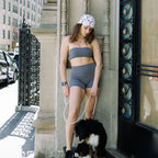 A woman wearing a matching gray two-piece set with a white patterned headband stands on a city sidewalk next to a black and white dog, holding a pearl leash.