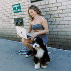 A woman in a gray strapless dress sits on a concrete sidewalk next to a brick wall, holding a silver laptop and petting a black and white dog. A blue leash lies on the ground, and a green sign on the wall reads 'SPRINKLERS IN GARAGE ONLY'.
