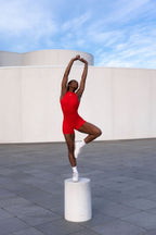 Model in sculpting red romper standing in ballet pose under blue sky — The Palm activewear