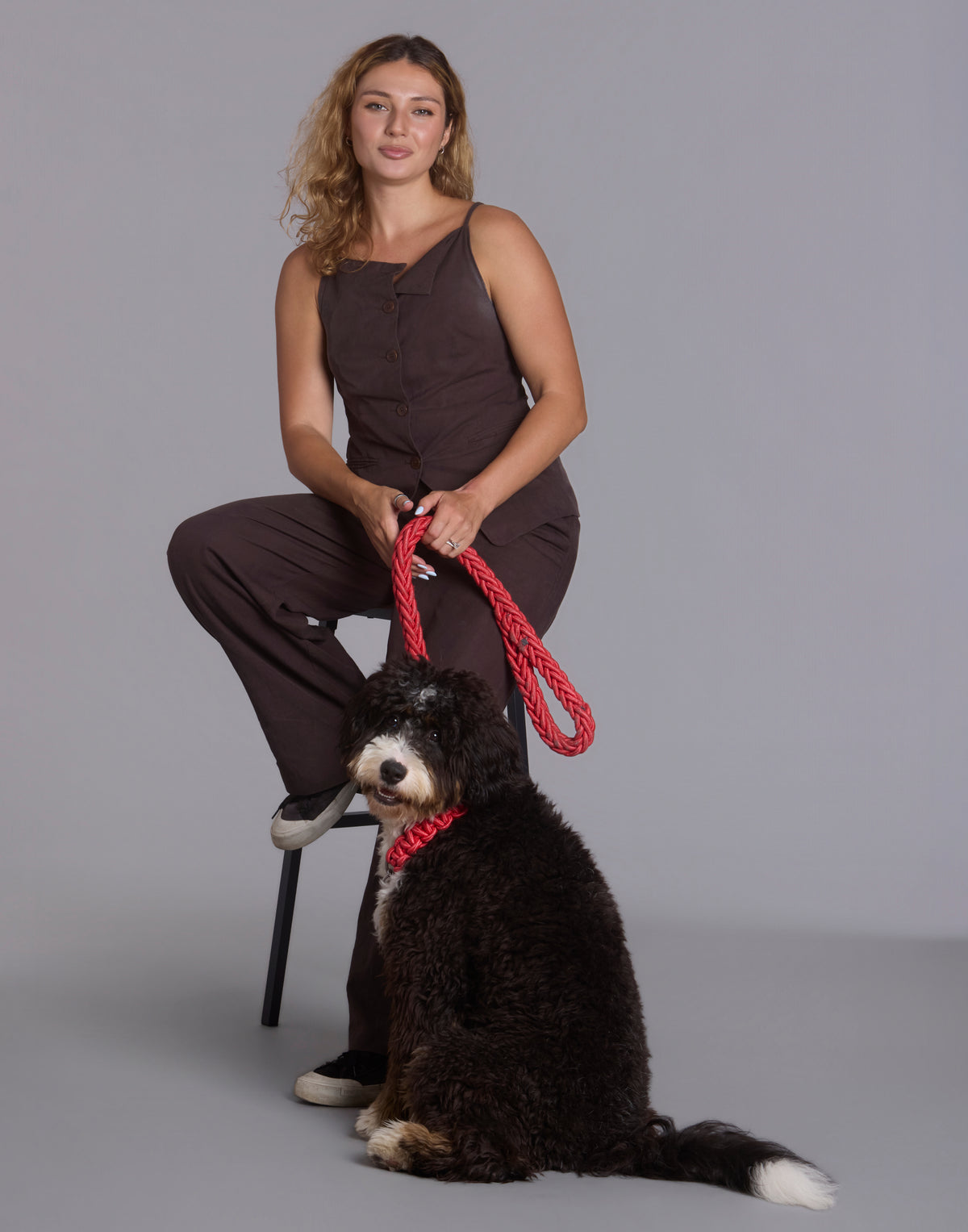 Full-length studio shot of woman sitting on chair holding red braided leash with fluffy black dog on the floor