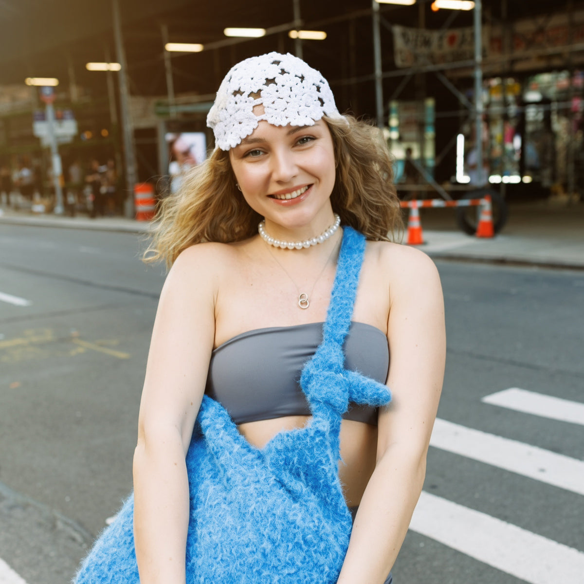 A woman standing on a city street crosswalk wearing a gray bandeau top and shorts, a white crochet cap, and holding a large fuzzy blue shoulder bag.