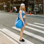 A woman stands on a city crosswalk wearing a gray sports bra and shorts, black sneakers, and a white patterned beanie, while carrying a large bright blue fuzzy shoulder bag. The background shows a city street with scaffolding and buildings.