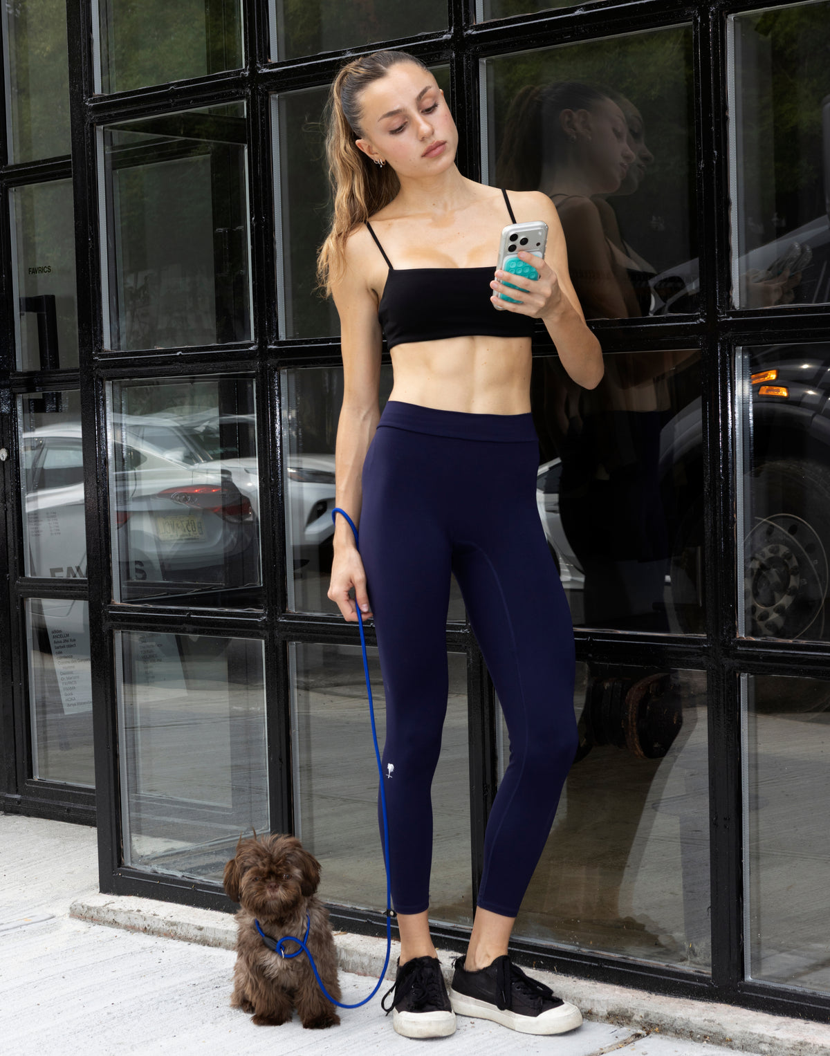 Front full-length view of woman holding smartphone standing with dog wearing high-waisted dark blue LiftLine sculpt leggings