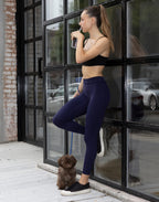 Sporty woman drinking from bottle standing with small dog wearing dark blue LiftLine sculpt leggings and black sports bra